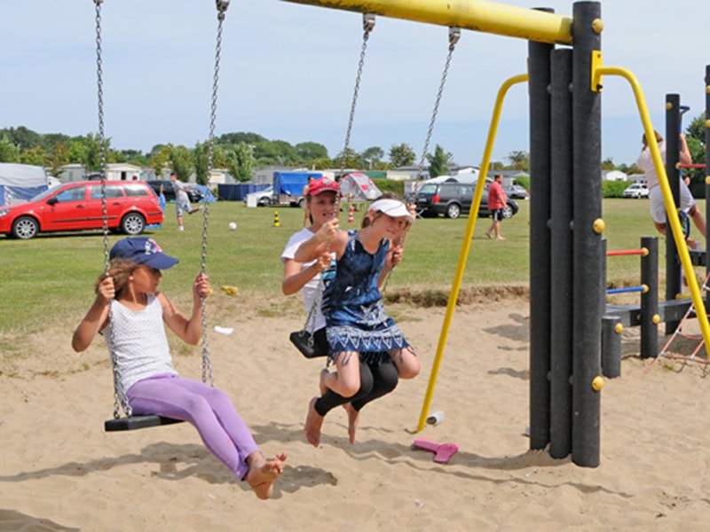 Gezellige speeltuin bij een stacaravan aan zee met kinderen die plezier maken op de schommel en speeltoren, ideaal voor familievakanties aan de kust.