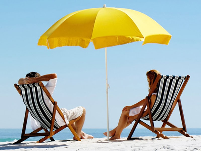 Gezicht op een strand met een blauwe lucht en zee, een paar zit onder een grote gele parasol op strandstoelen, vakantie, ontspanning, zon, zee, zeezicht, strandvakantie, aan zee, comfortabele accommodatie, perfecte strandvakantie.